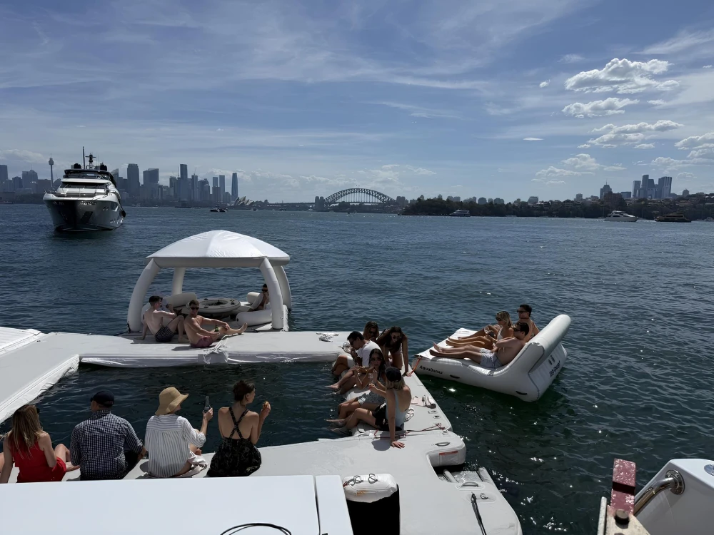 Guests relaxing on a floating platform beside a luxury yacht with Sydney Harbour, the Sydney Harbour Bridge and city skyline in the background.